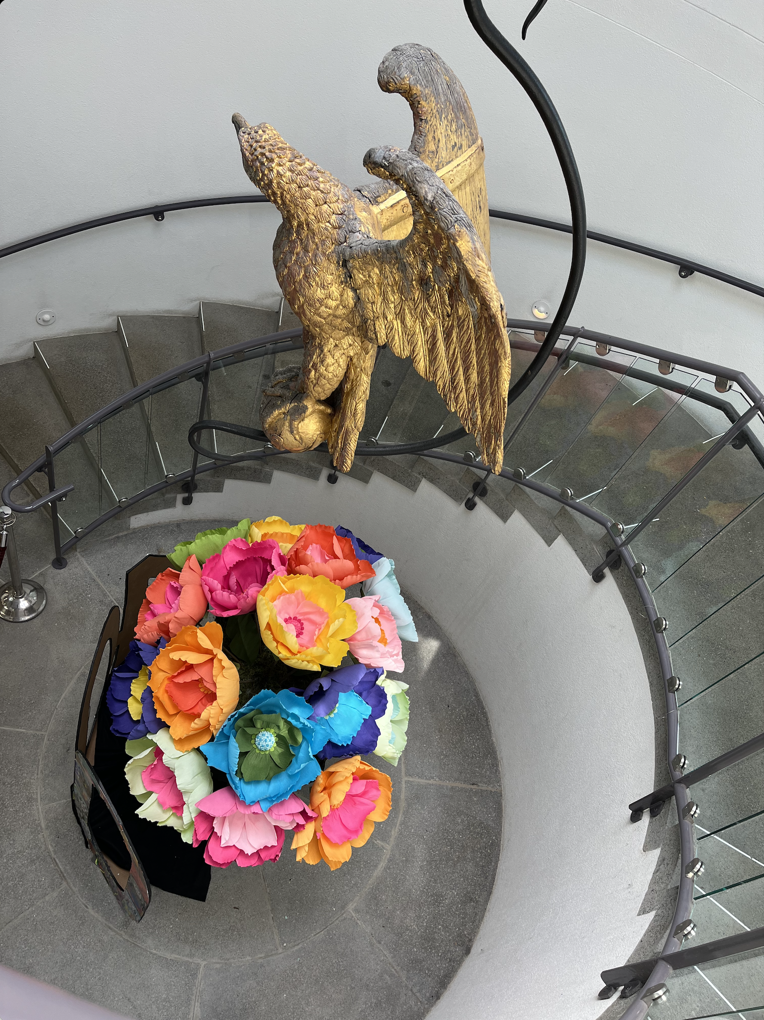 Perspective shot looking down at a circular stone staircase with a colorful flower arrangement at the bottom and a bronze eagle hanging above